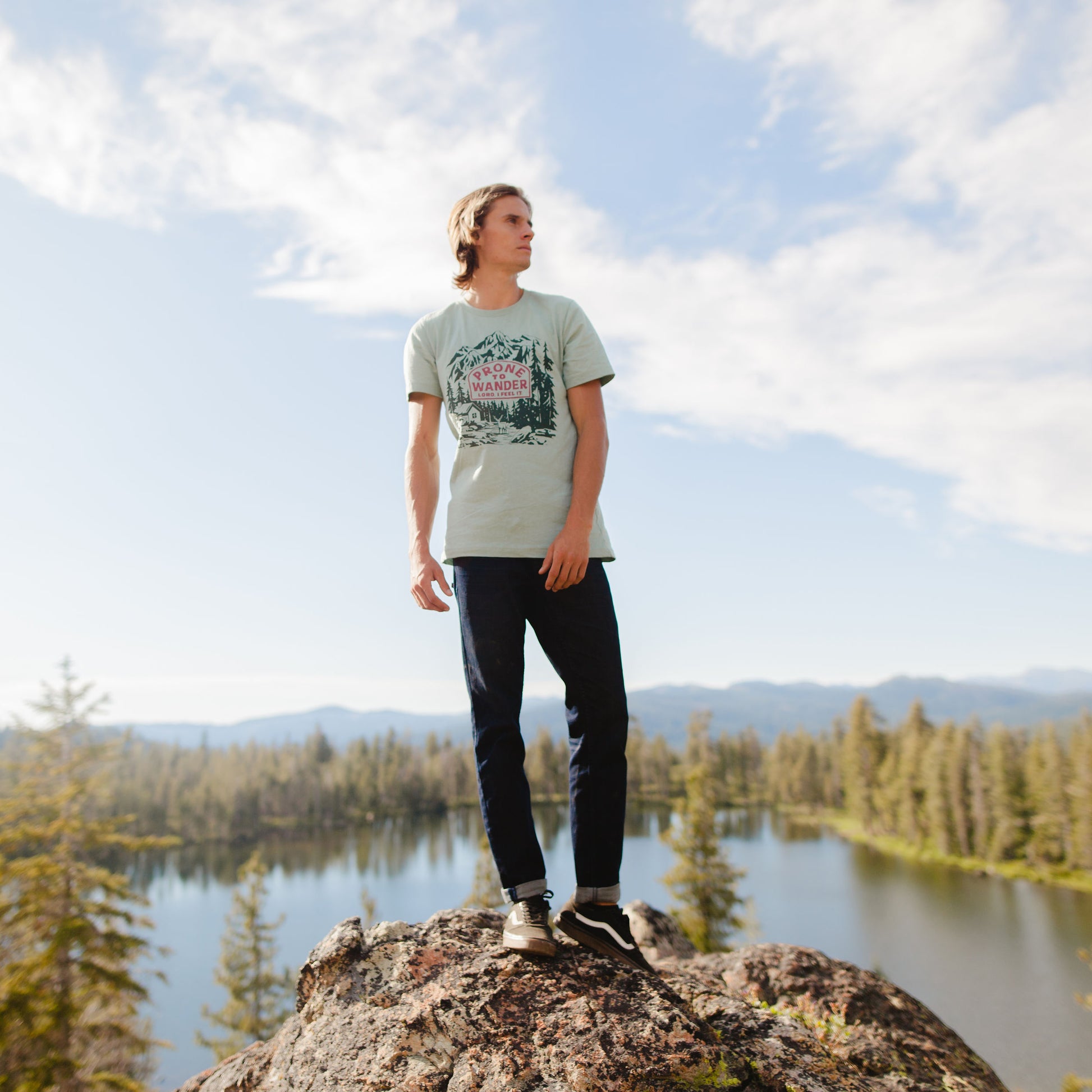 Young man wearing the Tugboat Jack Prone to Wander t-shirt while standing on a rock in the mountains with a lake in the background