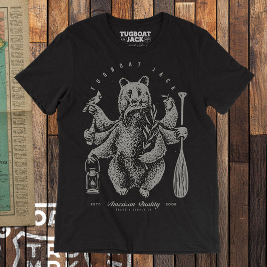 A black t-shirt on a wood floor. The shirt has an illustration of a six-armed bear holding various objects (a cardinal, a bottle of beer, a lantern, an apple, a paddle, and a willow brach). Above the bear is the headline "Tugboat Jack". Below the bear is the text "American Quality", "ESTD 2008", and "Trade & Supply".