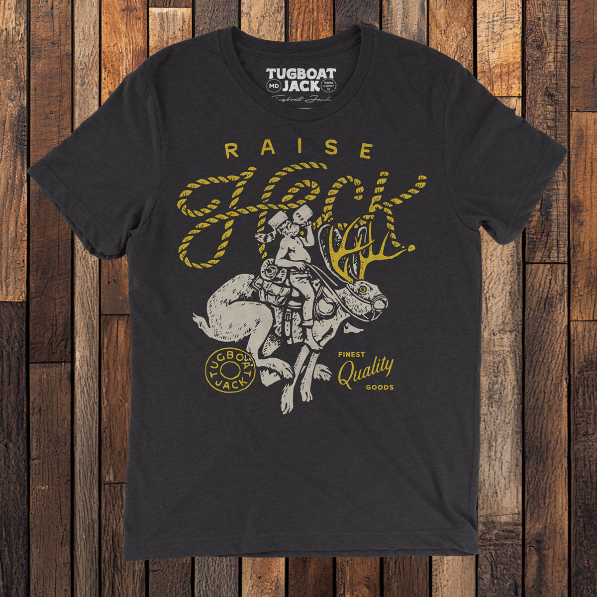 a photo of a black t-shirt on a wood floor. The shirt has a white and yellow illustration of a man riding a jackalope with the headline "Raise Heck" arched above. On either side of the illustration is the name "Tugboat Jack" and "Finest Quality Goods".