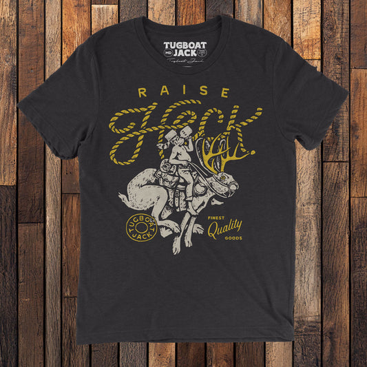 a photo of a black t-shirt on a wood floor. The shirt has a white and yellow illustration of a man riding a jackalope with the headline "Raise Heck" arched above. On either side of the illustration is the name "Tugboat Jack" and "Finest Quality Goods".