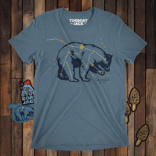 A grey-ish blue t-shirt laying on a wood floor with some small stickers around it. On the t-shirt is an illustration of a bear's silhouette. Overlayed on the illustration is the ursa major constellation. 