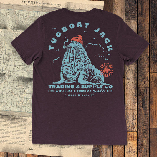A dark reddish color t-shirt laying on a wood floor and a paper star map. The illustration on the shirt is of a blue walrus wearing a red beanie and smoking a pipe. The text surrounding the shirt reads "Tugboat Jack", "Trading & Supply Co", "With just a pinch of salt", "Finest Quality", and "TRD MRK". There is a red stamp overlaying the design with a simplified walrus that reads "Seal of approval".